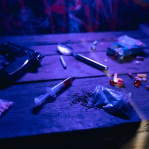 Wooden trunk with multiple types of drugs, a gun, syringe, teaspoon, cigarettes. Dark blue light.