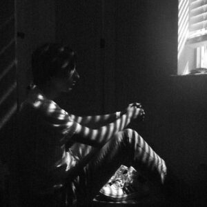 Man sitting down with back to wall looking out of window in dark room.